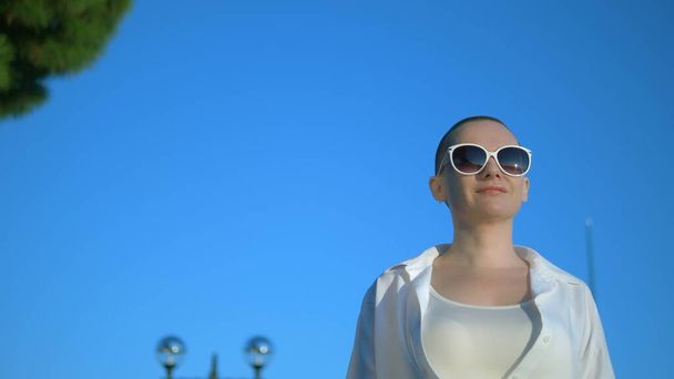 stylish bald girl in sunglasses and a white shirt walks the street against a blue sky and green trees - Fotografie, Obrázek