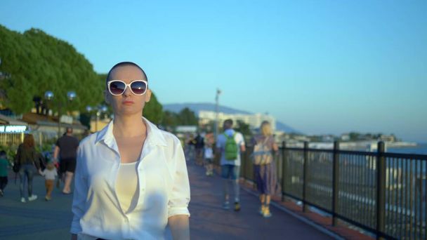 stylish bald girl in sunglasses and a white shirt walks the street against a blue sky and green trees - Fotoğraf, Görsel