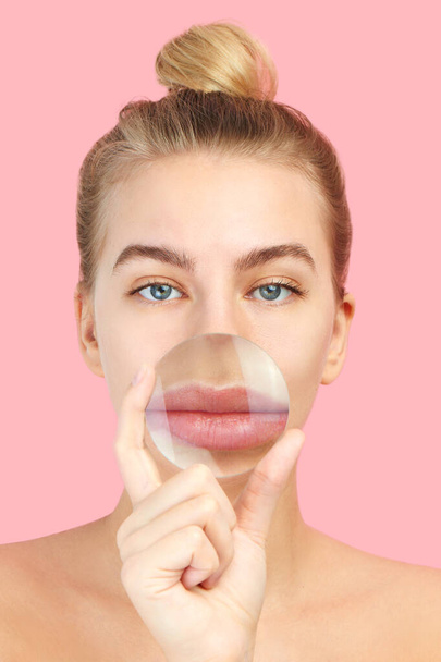 Beautiful young european woman with magnifying glass is about to do lip augmentation procedure close up on pink. Cosmetology, plastic surgery concept - Photo, Image