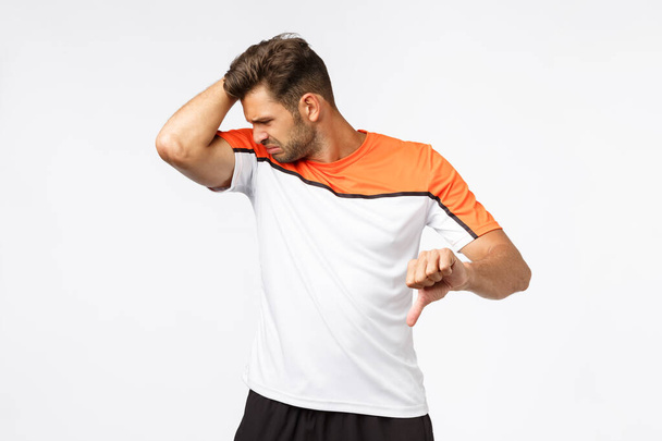 Guy feeling unconfident and upset as having problem with finding good deodorant. Masculine sportsman after workout raise arm and smell armpit, grimacing from disgust showing thumb-down displeased - Fotó, kép
