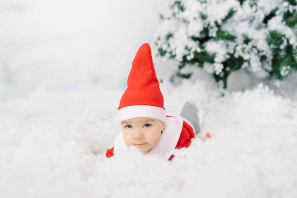Een vijf maanden oude baby in een kerstmankostuum ligt op kunstmatige sneeuw onder een kerstboom.. - Foto, afbeelding