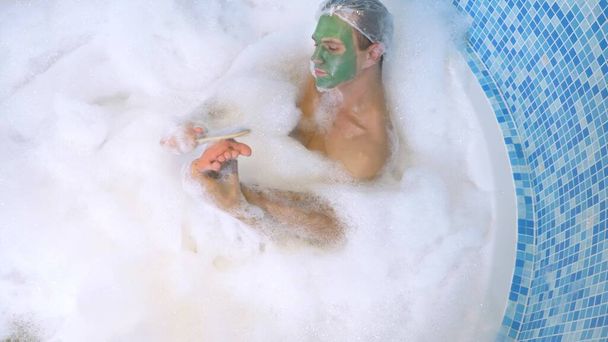 A man in a shower cap and a green face mask lies in a bath with copious foam and washes his foot with pumice. hygiene with scrub in the bath. view from above - Foto, afbeelding