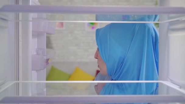young muslim woman in a national headscarf looks into an empty refrigerator and orders food at home through an app in her phone - Imágenes, Vídeo