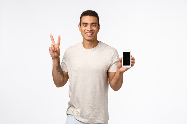 Friendly, cheerful handsome man in casual outfit, holding smartphone with blank screen, showing peace or victory sign, smiling, advertise site or app, recommend download mobile application - Photo, Image