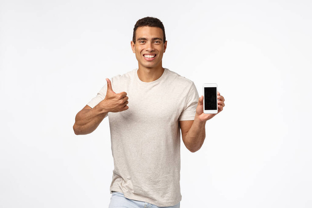 Satisfied, cheerful good-looking sexy young man in t-shirt, showing thumbs-up in approval or like gesture, smiling nod in agreement, recommend app, holding smartphone, introduce gadget - Fotoğraf, Görsel