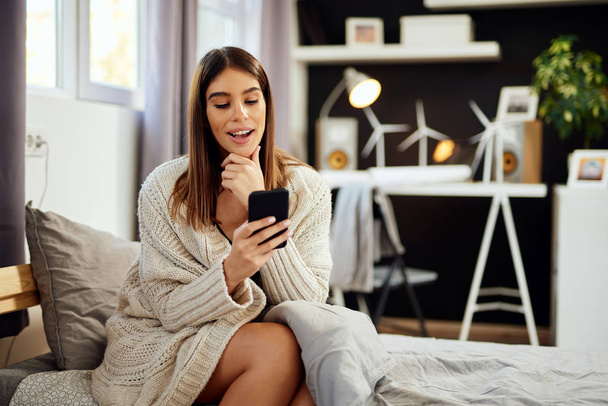 schöne lächelnde kaukasische Brünette sitzt in beigem Pullover auf dem Bett im Schlafzimmer sitzen und das Smartphone benutzen. - Foto, Bild