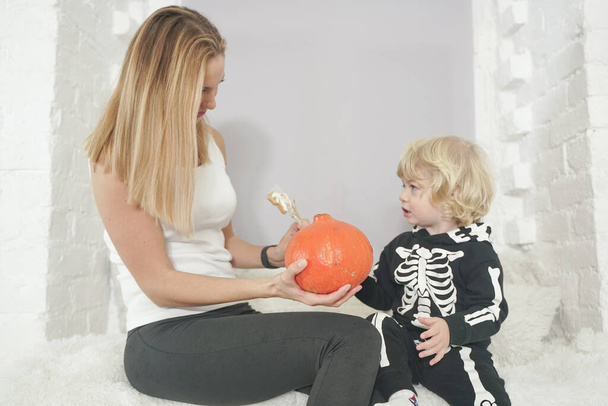 Młoda kobieta i jej syn mają jesień zabawy z dynią na białym parapecie, chłopiec ubrany w kostium szkielet - Zdjęcie, obraz