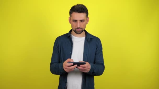 A young man in a blue shirt and white T-shirt is plays a game on a smartphone and loses, is upset. Yellow background with copy space. 4k slowmotion footage - Filmati, video
