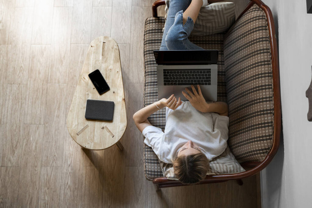 Woman with a laptop laying on a sofa. Study and work online, freelance. Self employed woman, girl working with her notebook laying on a couch with a phone, smartphone and ereader on table. - Photo, Image