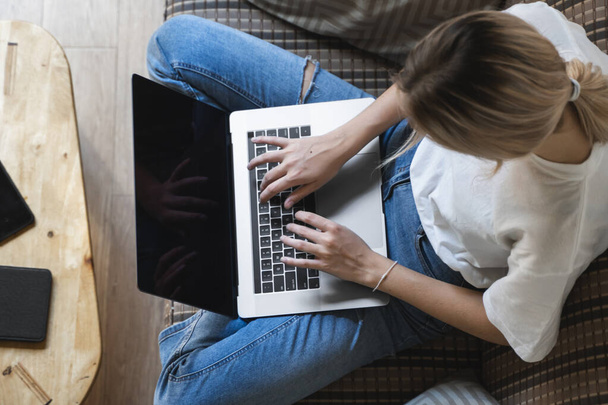 Woman with a laptop sitting on a sofa. Study and work online, freelance. Self employed woman, girl working with her notebook sitting on a couch with a phone, smartphone and ereader on table. - Photo, Image