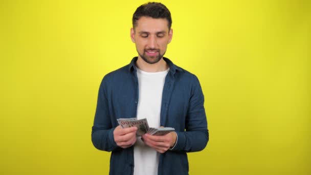 A young man in a blue shirt and white T-shirt is counting a pack of money and savour. Yellow background with copy space. 4k slowmotion footage - Кадры, видео