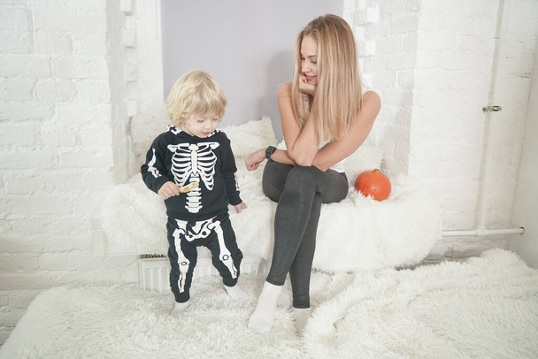 Jovem mulher e seu filho bebê se divertem no outono com abóbora no peitoril da janela branca, menino vestido com traje de esqueleto
 - Foto, Imagem