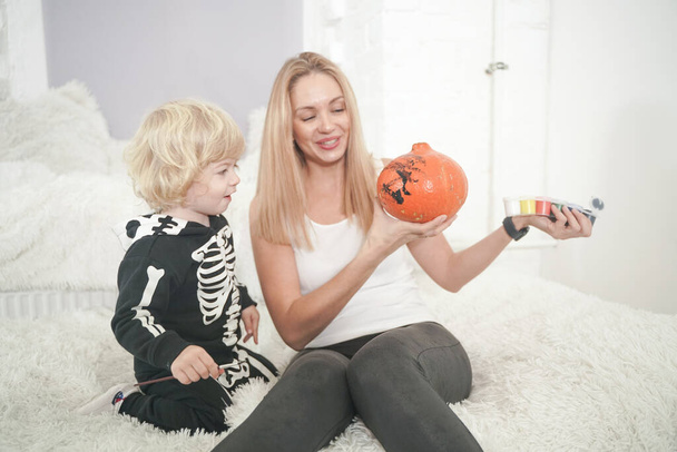 Młoda kobieta i jej syn mają jesień zabawy z dynią na białym parapecie, chłopiec ubrany w kostium szkielet - Zdjęcie, obraz