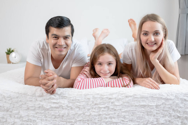 little girl smiling with mother and father on bed together in be - Zdjęcie, obraz