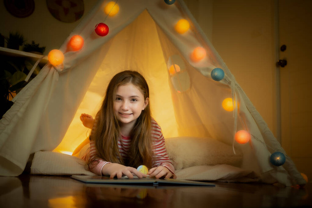 šťastná holčička čtení knihy a hrát kempování ve stanech doma - Fotografie, Obrázek