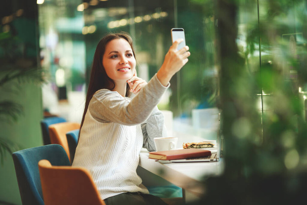 Portrait of young smiling girl make video call from smartphone to her best friend, having break, telling good news, works in cafe - Photo, Image