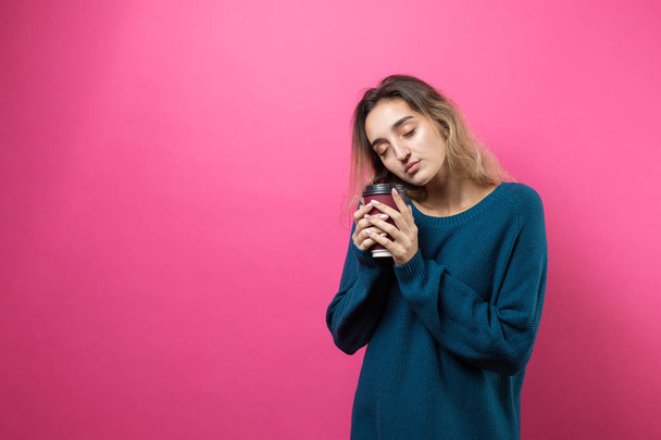 ピンクの背景にコーヒーを飲みながら青いセーターを着たグラマーな女性 - 写真・画像