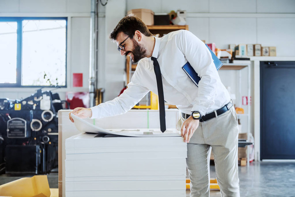 Smiling caucasian bearded graphic engineer with eyeglasses and in shirt and tie holding notebook under armpit and looking at printed sheets while standing in printing shop. - Photo, Image
