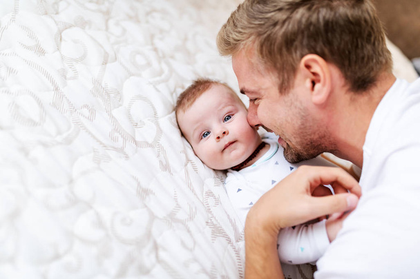 Nahaufnahme des schönen jungen kaukasischen Papas beim Kuscheln mit seinem sechs Monate alten Sohn. Baby liegt im Bett und umarmt Vater. - Foto, Bild
