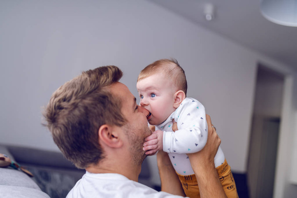 6 aylık beyaz bir çocuğun babasının burnunu ısırmasına yakın çekim yap. Yakışıklı genç baba kucağında bebek var. Diş büyütme kavramı. - Fotoğraf, Görsel
