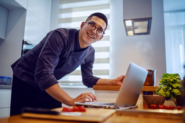 Lächelnder kaukasischer Koch in Uniform und mit Brille, der Rezepte auf dem Laptop betrachtet, während er in der Küche steht. - Foto, Bild