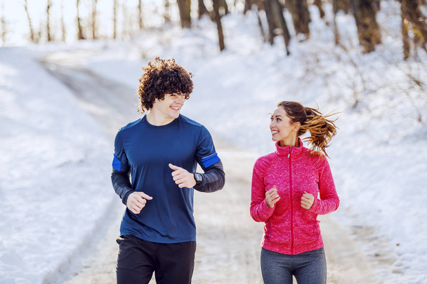 Feliz pareja caucásica sonriente en ropa deportiva corriendo en la naturaleza mientras se miran entre sí. Tiempo de invierno
. - Foto, imagen