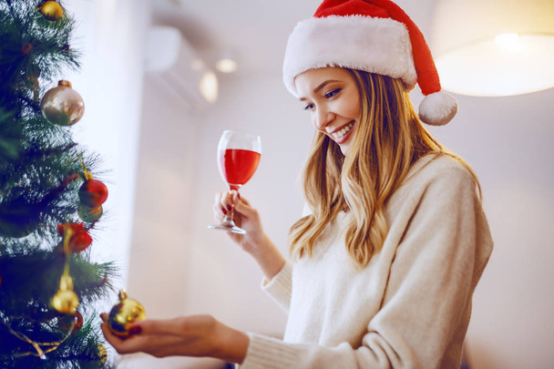 Hermosa mujer rubia caucásica sonriente con sombrero de santa en el ornamento conmovedor en el árbol de Navidad y beber vino mientras está de pie en la sala de estar
. - Foto, imagen