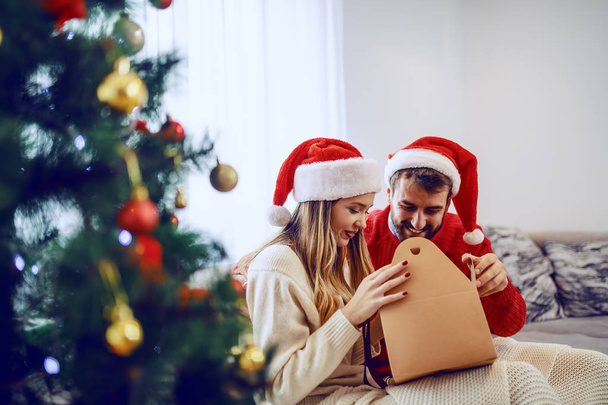 Adorable smiling caucasian couple with santa hats on heads siting on sofa in living room and opening gift. In foreground is christmas tree. - Fotografie, Obrázek