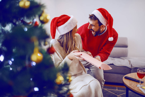 Amazed cute Caucasian blonde woman sitting on sofa in living room and receiving gift from her boyfriend. Both having santa hats on heads. In foreground is christmas tree. Living room interior. - Foto, Imagem