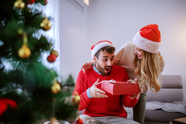 Charming caucasian woman giving christmas gift to her boyfriend. Both are dressed in sweaters and having santa hats on heads. Living room interior, christmas holiday concept. - Foto, imagen