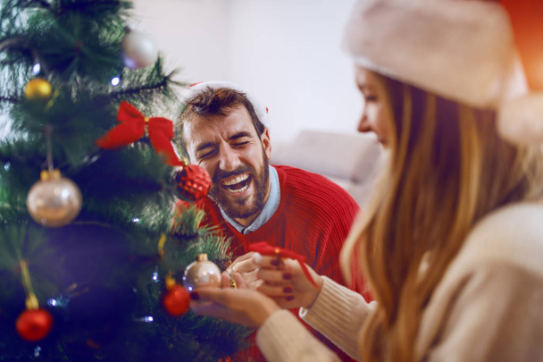 Riéndose guapo barbudo caucásico hombre ayudando a su novia a decorar el árbol de Navidad. Ambos tienen sombreros de Santa Claus en la cabeza. Salón interior
. - Foto, imagen