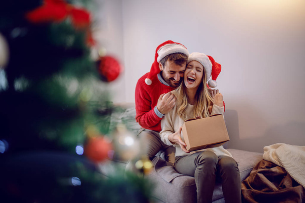 Podekscytowana piękna biała młoda kobieta trzymająca prezent świąteczny i dziękująca swojemu kochającemu chłopakowi. Mężczyzna przytulający kobietę. Oboje mają czapkę Mikołaja na głowach. Na pierwszym planie jest choinka. - Zdjęcie, obraz