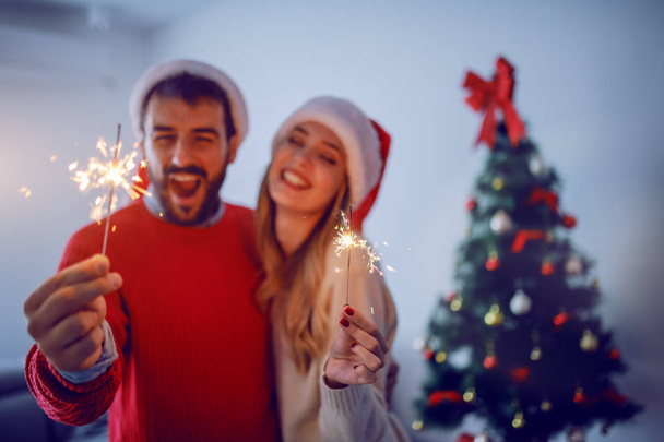 Ευτυχισμένο ζευγάρι Καυκάσιων με καπέλα Σάντα σε κεφάλια που κρατάνε ψεκαστήρες ενώ αγκαλιάζονται και στέκονται στο σαλόνι. Στο βάθος είναι το χριστουγεννιάτικο δέντρο. Επιλεκτική εστίαση στους ψεκαστήρες. - Φωτογραφία, εικόνα