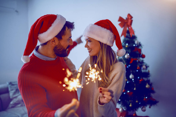 glückliches kaukasisches attraktives Paar, das sich gegenseitig zum neuen Jahr gratuliert. Paar mit Weihnachtsmützen auf dem Kopf und Sprinklern im Wohnzimmer. Im Hintergrund steht der Weihnachtsbaum. - Foto, Bild