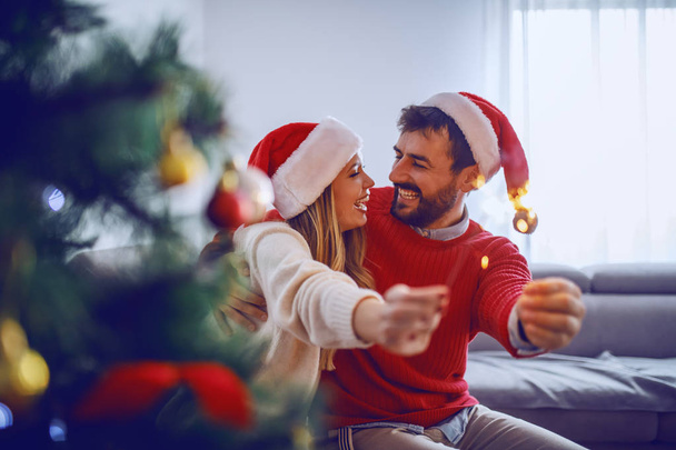 glückliches kaukasisches Paar mit Weihnachtsmützen auf dem Kopf, das auf dem Sofa sitzt, Sprinkler in der Hand und Neujahr feiert. Im Vordergrund steht der Weihnachtsbaum. - Foto, Bild