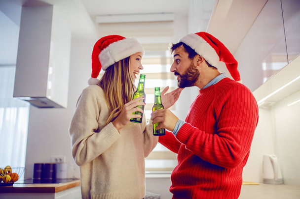 Side view of cute young caucasian couple with santa hats on heads holding beer and talking while standing in kitchen. Christmas time. - Photo, image