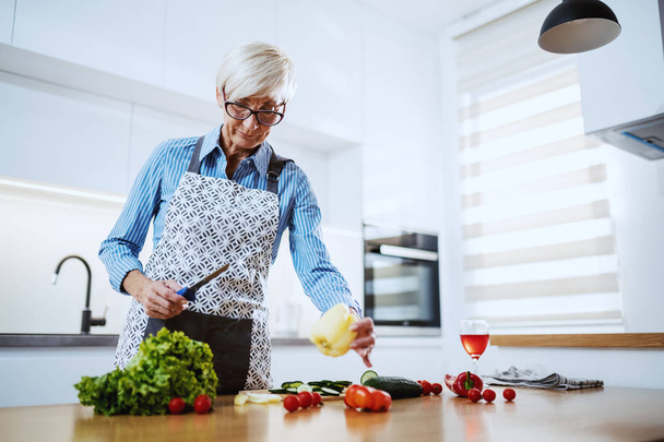 schöne kaukasische blonde Seniorin in Schürze, die in der Küche steht und gesunde Mahlzeiten zubereitet. Auf dem Küchentisch liegen Gemüse und ein Glas Wein. - Foto, Bild