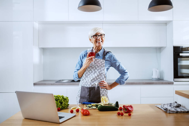 Femme âgée caucasienne souriante dans un tablier debout dans la cuisine, buvant du vin et préparant un dîner sain. Sur le comptoir de la cuisine sont poivrons, laitue, tomates cerises et ordinateur portable
. - Photo, image