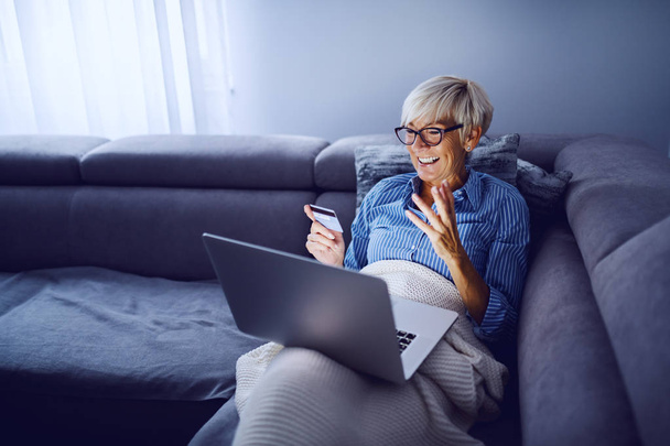 Veselý okouzlující kavkazský senior blond žena s krátkými vlasy a brýlemi sedí na pohovce v obývacím pokoji, drží notebook v klíně a pomocí kreditní karty pro on-line nakupování. - Fotografie, Obrázek
