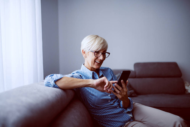 Vonzó mosolygós fehér szőke idős nő rövid hajjal ül a kanapén a nappaliban ablak mellett, és okos telefont használ. - Fotó, kép