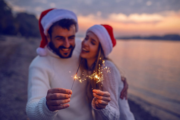 überglückliches kaukasisches verliebtes Paar, das an der Küste in Flussnähe steht und Wunderkerzen in der Hand hält. Beide tragen weiße Pullover und Weihnachtsmützen auf dem Kopf. Selektiver Fokus auf Wunderkerzen. - Foto, Bild