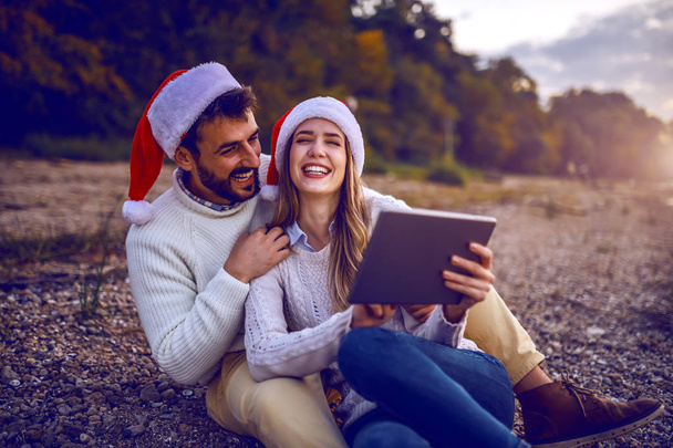 Nehir kıyısında oturup tablet kullanan mutlu, beyaz, modaya uygun genç bir çift. Kadın tableti tutarken, erkek kadına sarılıyor. İkisinin de kafasında Noel Baba şapkası var.. - Fotoğraf, Görsel