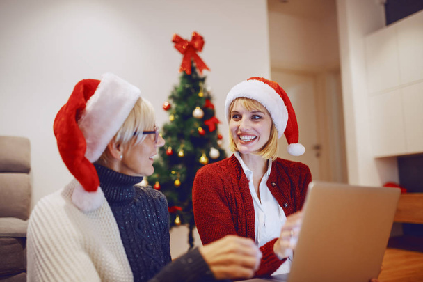 Gyönyörű fehér szőke nő ül az anyjával a nappaliban és laptopot használ. Mindkettőnek télapó sapka van a fején. A háttérben díszített karácsonyfa. - Fotó, kép