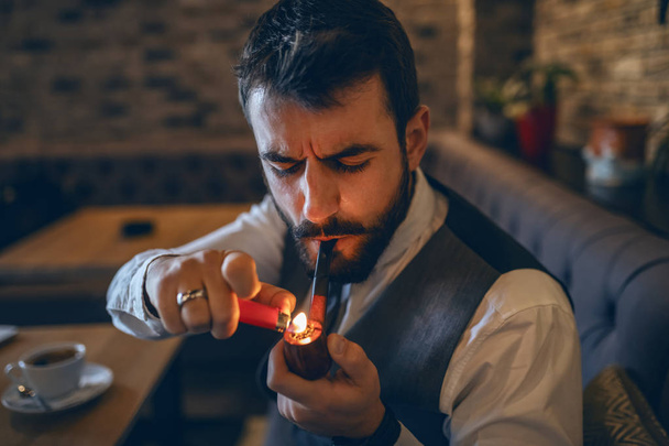 Sofisticado hombre de negocios barbudo caucásico encendiendo una tubería mientras está sentado en la cafetería
. - Foto, imagen
