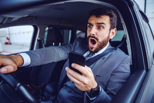 Homem de negócios caucasiano incrível em terno carro de condução e usando telefone inteligente ao mesmo tempo
. - Foto, Imagem