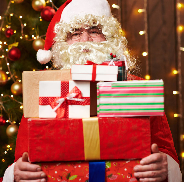 Weihnachtsmann posiert mit vielen Geschenken, sitzt drinnen neben geschmücktem Weihnachtsbaum mit Lichtern - frohe Weihnachten und frohe Feiertage! - Foto, Bild