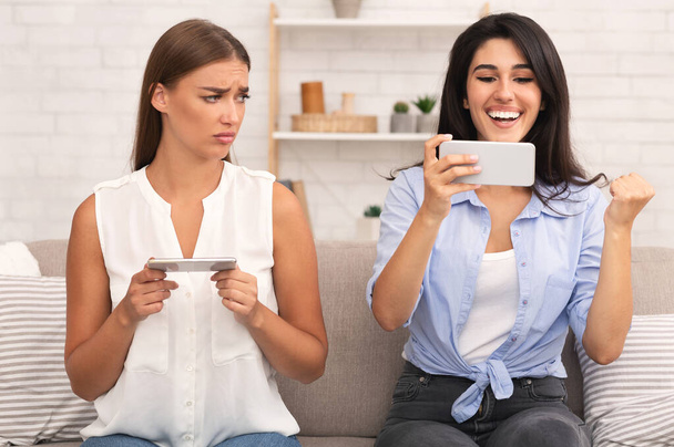 Mädchen verliert Handyspiel, wenn sie mit Freundin drinnen auf Couch sitzt - Foto, Bild