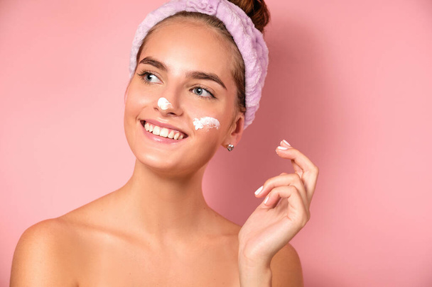 A beautiful girl with a bandage on her head a pink background smiles with smears of cream on her face - Foto, Imagen