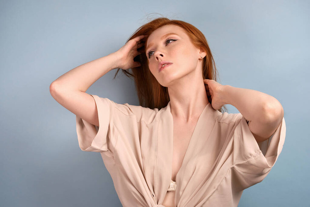 A red-haired girl in a light robe stands on a blue background and lifts her chin, looks to the side, straightening her hair. - Fotoğraf, Görsel