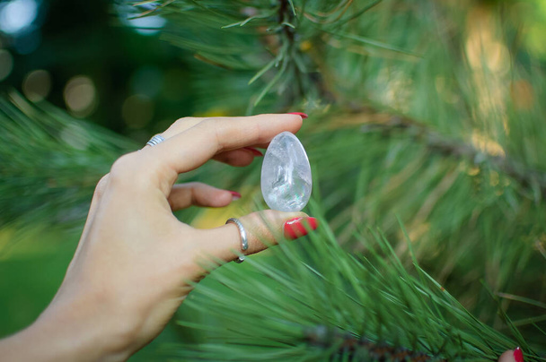 Frauenhand mit transparentem Amethyst-Quarz-Yoni-Ei für Vumfit, Imbuilding oder Meditation. glänzende Kristall-Ei in den Händen auf Kiefernzweigen Hintergrund im Freien. Frauengesundheitskonzept - Foto, Bild